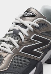 Κοντινό πλάνο του αθλητικού παπουτσιού New Balance 9060 σε γκρι και ασημί, με λευκά κορδόνια και λεπτομέρειες από δίχτυ στο πάνω μέρος.