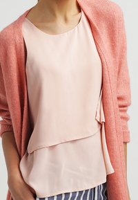 Jasnoróżowa warstwowa bluzka z okrągłym dekoltem pod miękkim koralowym kardiganem w prążki. Widoczne spodnie w paski w kolorze granatowym i białym.