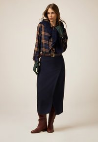 Karierte Bluse in Marineblau und Brauntönen, kombiniert mit einem marineblauen Strickschal, dazu ein marineblauer Rock und braune kniehohe Stiefel.