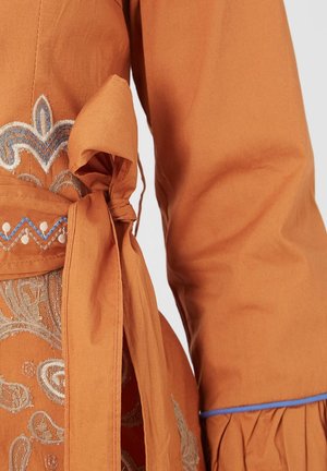 La tela de algodón naranja presenta un detallado bordado en beige y azul. Un cinturón atado añade forma, con un volado texturizado en el puño.