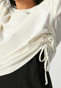 Top bianco realizzato in morbido tessuto con dettaglio arricciato sui lati e cordini. Marchio discreto vicino al collo. Sfondo nero.