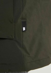 Zielona, lekka kurtka o gładkiej fakturze, z boczną kieszenią i małą czarno-białą metką marki na dole.