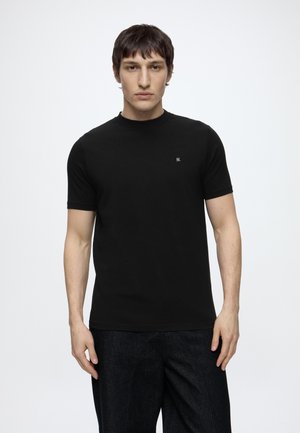 Modèle masculin debout, face à l’avant, portant un t-shirt noir uni à manches courtes et un pantalon foncé sur fond blanc.
