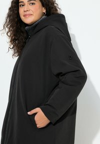 Sort hættejakke med tekstureret stof, frontlynlås og sidelommer. Har en logo-accent på venstre ærme.