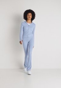 Tuta aderente zipato di colore azzurro chiaro, con maniche lunghe e pantaloni svasati; texture liscia, dettagli minimi, abbinato a sneaker bianche.