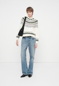 Oseba v belem puloverju z vzorcem, modrih kavbojkah, črnih špicastih čevljih in črni torbi čez ramo, stoji ob belem ozadju.