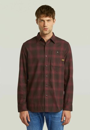 Hombre con una camisa de cuadros en rojo oscuro y negro abotonada, con bolsillo en el pecho y jeans azules, de pie contra un fondo claro y liso.