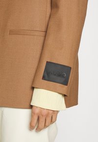 Brauner Blazer aus strukturiertem Stoff, mit einem schwarzen Patch mit geprägtem "HUGO"-Logo und rotem Stitching, getragen über einem hellen Hemd.