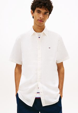 Jonge man met krullend haar, gekleed in een wit overhemd met korte mouwen over een gestreept T-shirt en marineblauwe korte broek, handen in de zakken.