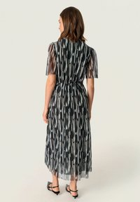 Mustakankaiset midi-mekko, jossa on läpikuultavat hihat, kerrottu vyötärö ja lentävä rakenne. Korostettu minimalistisilla mustilla kengillä.
