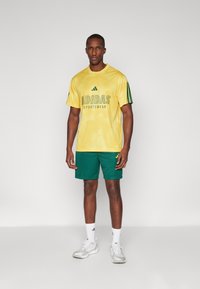 Rumena mrežasta športna majica z zelenimi detajli in logotipom "ADIDAS SPORTSWEAR", v kombinaciji z zelenimi kratkimi hlačami z teksturiranim vzorcem.