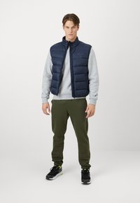 Marineblauer Steppweste über einem grauen Sweatshirt, kombiniert mit olivgrünen Jogginghosen. Schwarze Sneakers mit gelben Akzenten. Sauberes und minimalistisches Design.