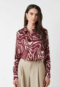 Woman with long dark hair wearing silver hoop earrings, a maroon and beige abstract patterned button-up blouse, and beige high-waist pants.
