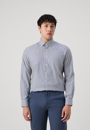Gestreiftes Button-Down-Hemd in Marineblau und Weiß, mit einem Umlegekragen und langen Ärmeln. Kombiniert mit maßgeschneiderten blauen Hosen für einen formellen Look.