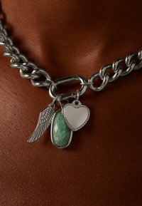 Chunky zilveren ketting met drie bedels: een vleugel, een groene steen en een hartvormige hanger, allemaal met ingewikkelde details.