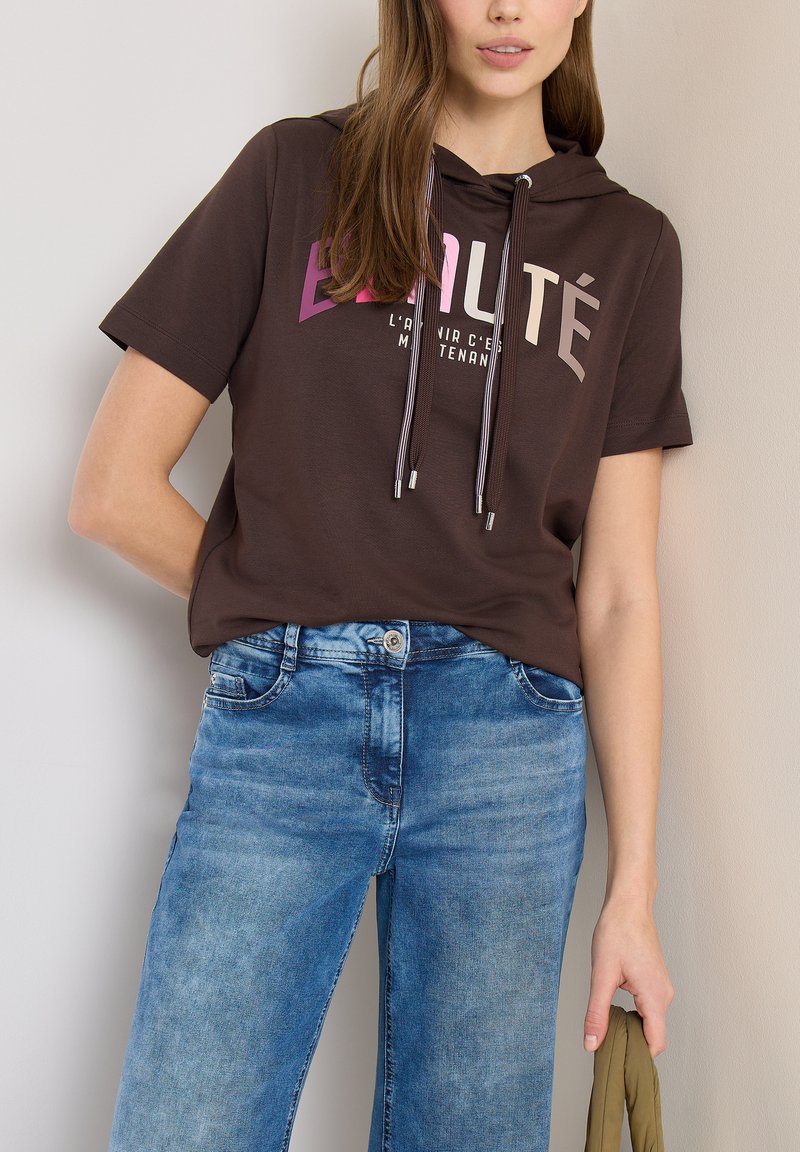 Jeune personne portant un sweat à capuche marron à manches courtes avec le texte « Beauté » et un jean bleu, tenant un tissu vert olive dans la main droite.