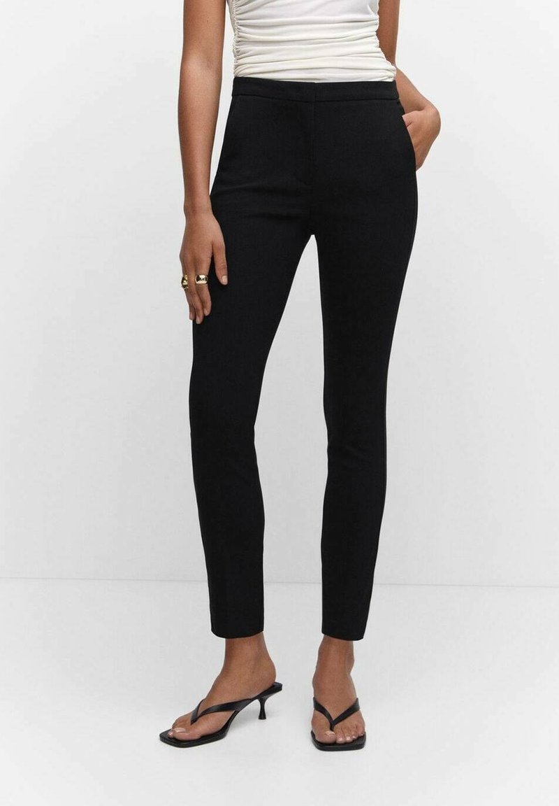 Pantalon noir slim porté avec des sandales à talons noires et un top blanc froncé sans manches, une main dans la poche et des bagues aux doigts.