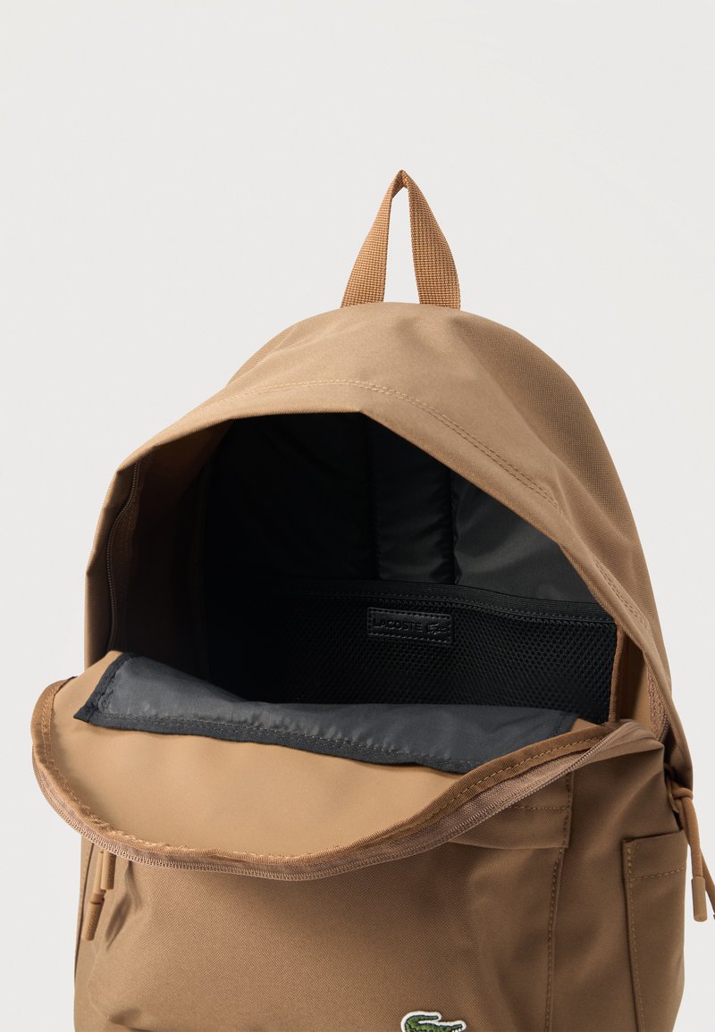 Zaino beige con scomparto principale aperto che mostra una tasca interna in rete nera e una maniglia a anello in tessuto su uno sfondo chiaro.