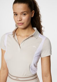 Ljusbeige pikétröja med korta ärmar, har vita sidopaneler och en liten logotyp på bröstet. Slät tygkvalitet.