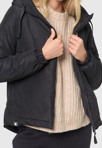 Sort puffjakke med lynlås foran, hætte og elastiske manchetter, lagdelt over en beige strikket sweater med et struktureret mønster.