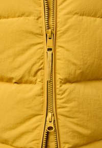Giacca isolante gialla con zip anteriore. Presenta una texture liscia e un design gonfiabile. Il tirazip è marchiato. Lo sfondo è semplice.