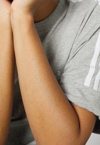 Grå t-shirt med kort ärm och vita st stripes på axlarna. Hudtonen är synlig med små texturdetailleringar på armarna.