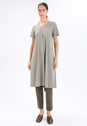 Donna che indossa una tunica ampia e a maniche corte di colore grigio chiaro sopra pantaloni verde oliva e scarpe marroni senza lacci, in piedi su uno sfondo bianco semplice.