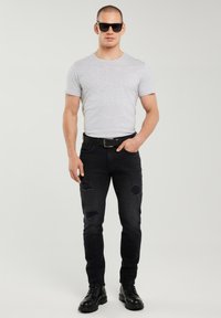 Mand iført sorte solbriller, lysegrå t-shirt, sorte revnede jeans, sort bælte og sorte støvler, stående med den ene hånd i lommen.