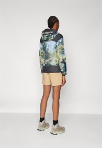 Casaco Columbia leve e multicolorido com estampado de natureza, detalhes em preto e capuz com cordão. Usado sobre calções bege e botas de caminhada.