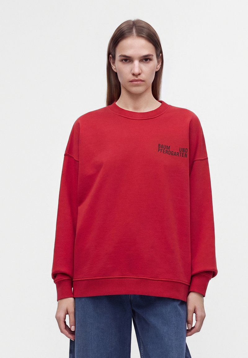 Jeune personne aux cheveux bruns et raides porte un sweat-shirt rouge oversize avec un texte noir sur la poitrine gauche et un jean bleu, debout devant un fond blanc.