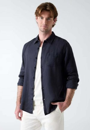 Joven con cabello castaño claro que lleva una camisa negra abotonada sobre una camiseta blanca y pantalones blancos, de pie frente a un fondo liso.