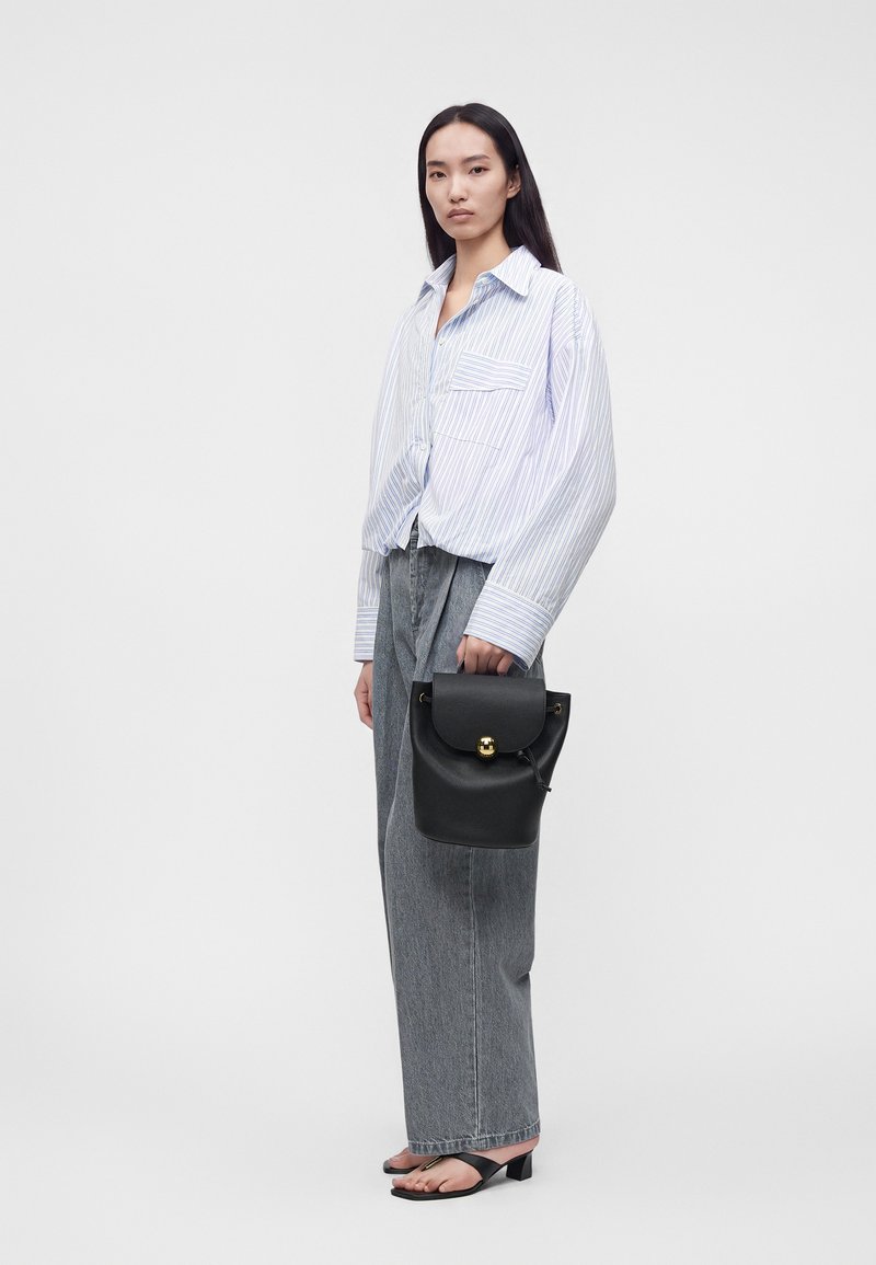 Femme portant une chemise rayée ample, jean gris à jambes larges, sandales noires à talons, tenant un petit sac à main noir avec une boucle dorée.