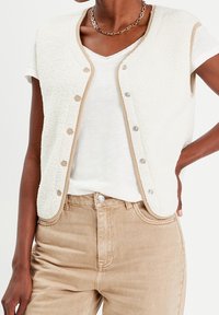 Vit fleeceväst med beige detaljer, med tryckknappar. Buren över en vit T-shirt och beige byxor, som visar en strukturerad yta.