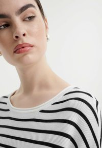 Gestreepte trui met zwart-witte horizontale lijnen, een ronde halslijn en een gestructureerde stof. Model toont een neutrale expressie.