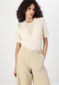 Crème-kleurige gebreide top met korte mouwen, voorzien van een gestructureerd patroon. Gecombineerd met hoog-getailleerde beige broeken met een enkele knoopdetails.