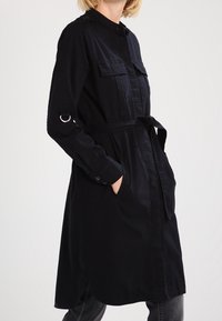 Kobieta w długim czarnym płaszczu z paskiem, z kieszeniami na piersi i bocznymi, stoi z jedną ręką w kieszeni.