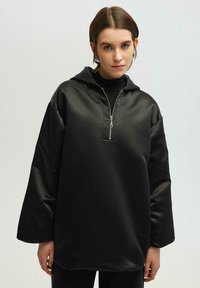 Felpa con cappuccio nera realizzata in un tessuto liscio e lucido. Presenta una mezza cerniera sul fronte e maniche ampie, con una silhouette dal taglio rilassato.
