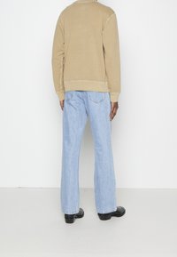 Beige huvtröja med ribbad fåll, bärs över ljusblå vida jeans. Svarta skor synliga. Bakifrån mot en enkel vit bakgrund.