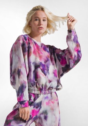 Blondyna kobieta w luźnej, kolorowej bluzie tie-dye i pasujących spodniach, trzymająca pasemko włosów na jednolitym białym tle.