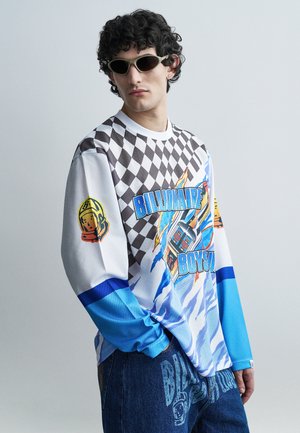 Ung mand med krøllet hår iført mørke solbriller, en farverig langærmet trøje med grafisk tryk og teksten "Billionaire Boys" samt blå jeans med print.