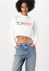 Fehér, rövidített pulóver, amelyen a "TOMMY" kék és piros hímzéssel szerepel, kerek nyakkivágással és bordás mandzsettákkal, világos kék, magas derekú farmernadrággal párosítva.