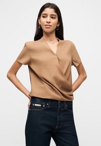 Καφέ μπλουζάκι με κοντό μανίκι και σχέδιο τύπου wrap, με ομαλή υφή και διακριτική λάμψη, συνδυασμένο με σκούρο τζιν τζιν με ετικέτα.