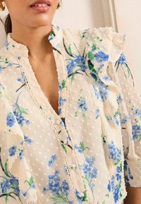 Blumenbluse mit einem Rüschensaum am Kragen und an den Ärmeln, aus leichtem, cremefarbenem Stoff mit blauen Blumendrucken und strukturierten Punktelementen.