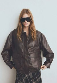 Chaqueta de cuero marrón con cremallera frontal, dos grandes bolsillos y dobladillo elástico. El modelo lleva gafas de sol negras de gran tamaño. Acabado texturizado.