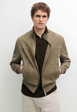 Mężczyzna ubrany w jasnobrązową zamszową kurtkę na ciemnobrązowej koszuli, z rękami w kieszeniach kurtki, stojący na jednolitym białym tle.