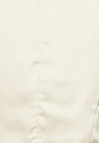 Krémová látka s hladkým, mírně lesklým povrchem. Obsahuje jemné švy a přiléhavý střih. Žádné viditelné vzory nebo kovové prvky.