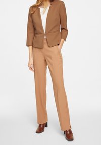 Femme portant un blazer camel et un pantalon droit taille haute assorti avec des bottes à talons marron, debout les mains dans les poches sur fond blanc.