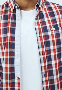 Karierte Hemd in Rot, Weiß und Blau mit einem hellblauen Kragen und einer Brusttasche. Darunter ist ein schlichtes weißes T-Shirt sichtbar.