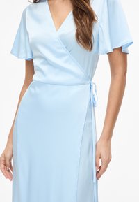 Femme portant une robe portefeuille bleu clair avec des manches courtes volantées nouée à la taille, debout devant un fond blanc.