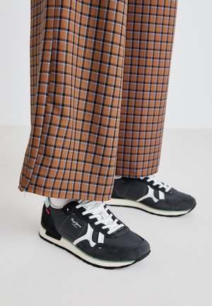 Sorte og grå sneakers med hvide accenter, der har en blanding af ruskind og mesh-materialer, parret med brune ternede vide bukser.